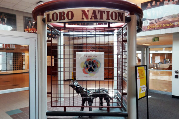 The entrance features a sculpture of a wolf displayed within a red, cylindrical cage-like structure with "Lobo Nation" written on the top. The background shows hallways and display cases filled with various items, likely in a school setting.