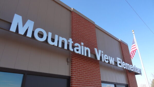 A building with a sign that reads "Mountain View Elementary" in large white letters. The structure features red brick and beige paneling. An American flag is visible on the right side of the image against a clear blue sky.