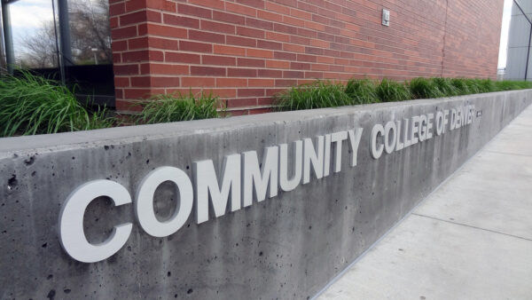 A concrete wall with large white letters reads "Community College of Denver." The wall is located next to a sidewalk and in front of a brick building with tall green plants along the top edge.