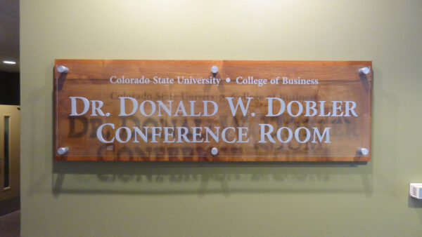 A wooden sign with glass panel affixed by metal pins, mounted on a green wall reads "Dr. Donald W. Dobler Conference Room" with the subheading "Colorado State University - College of Business" engraved at the top.