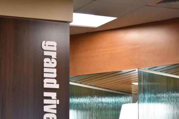 A modern office workspace inside Grand River Health. The office features a wooden partition with "grand river Health" written vertically in white letters. Behind the partition, there are desks with computers and chairs, and a glass wall with vertical lines.