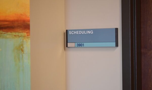 A sign on an office wall reads "SCHEDULING" with the room number "2901" below it. The wall has a modern, abstract painting with warm tones on the left side.