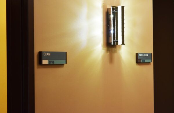 A beige wall with a modern wall sconce emitting light. Two signs are visible: one on the left reads "EXIT" in uppercase letters, and the one on the right reads "ELEVATOR" in uppercase letters. Both signs are green with white text.