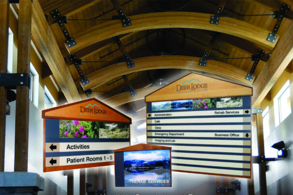 Wooden directional signs with text and images are displayed inside a building with a wooden arched ceiling and metal beams. The signs indicate locations for "Activities," "Patient Rooms 1-1," and various hospital services. Text at the bottom reads "Hospital - Deer Lodge, MT.