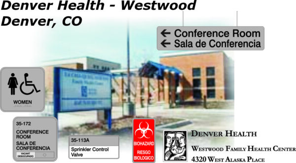Image of Denver Health - Westwood Family Health Center in Denver, CO. The foreground shows various signs including directions to the conference room in both English and Spanish, a women's restroom sign, sprinkler control valve, and a biohazard warning.