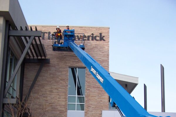 A blue Genie S-60 boom lift is extended to allow two workers to work on signage for the building.
