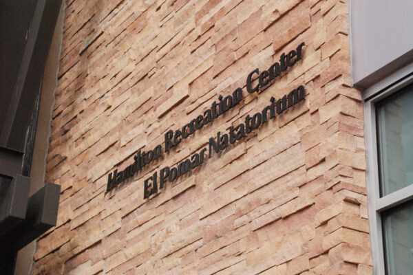 A sign mounted on a textured brick wall reads "Juan Tabo Recreation Center El Pinar Natatorium." The sign is composed of black letters and located near a corner of the building with a window partially visible to the right.