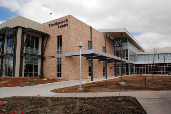 A brick and glass building named "The Maverick Center" with large windows and a modern design is shown. The entrance features an overhang and a paved pathway, surrounded by a lawn and landscaping under a partly cloudy sky.