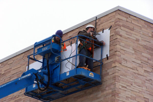 Two workers in safety gear are elevated on a blue Genie S-60 boom lift, performing maintenance or construction on the exterior sign on a brick building.
