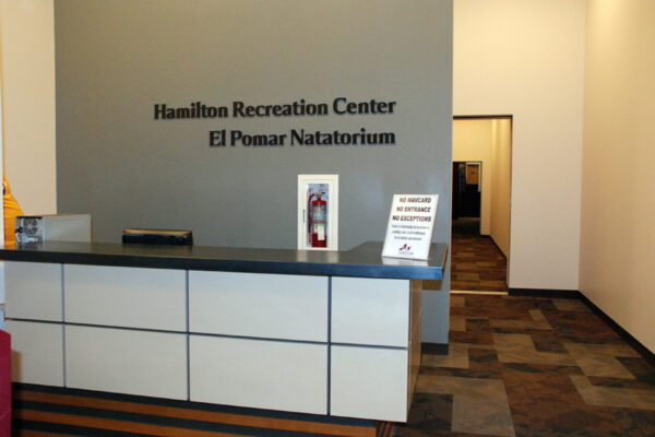 A reception desk at Hamilton Recreation Center with a sign that reads "Hamilton Recreation Center El Pomar Natatorium" on the wall behind it. A sign on the desk reads "No Swimming If a Lifeguard Is Not Present." A hallway extends to the right.