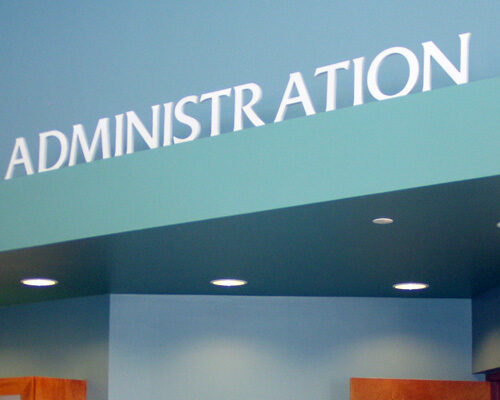 A teal-colored wall with the word "ADMINISTRATION" in large white letters at the top. There is a partially open wooden door with a glass window panel to the right and a wooden display case to the left. The area is well-lit with recessed ceiling lights.