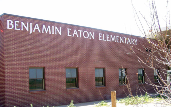 A brick building with the words "Benjamin Eaton Elementary" displayed prominently on the front. The building has several windows and minimal landscaping, with some plants and a tree visible in the foreground. The sky is clear and sunny.