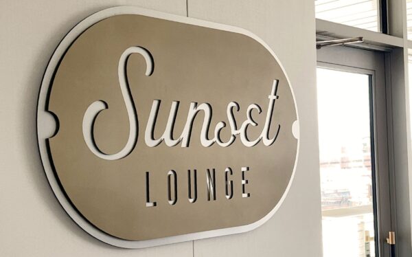 A metallic sign on a wall reads "Sunset Lounge" in elegant script. The background of the sign is a reflective silver color. To the right, there is a window or door with glass panes, allowing light to enter the space.