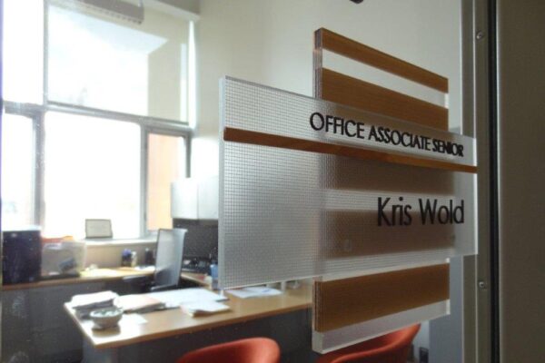 Office door with a nameplate reading "Office Associate Senior, Kris Wold." The door is partially open, revealing a well-lit office with a desk cluttered with papers and a laptop, and two red chairs in front of it. The room appears bright with sunlight coming through windows.