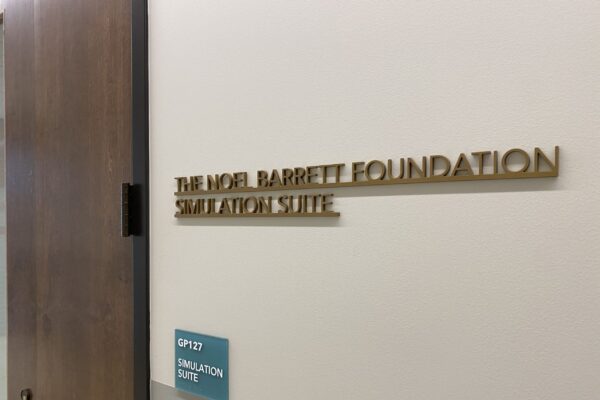 A door sign reads "THE NOEL BARRETT FOUNDATION SIMULATION SUITE" in raised gold letters. Below the sign is a blue and white room placard with the text "CP 127 SIMULATION SUITE." The door is slightly open, revealing a part of the hallway.