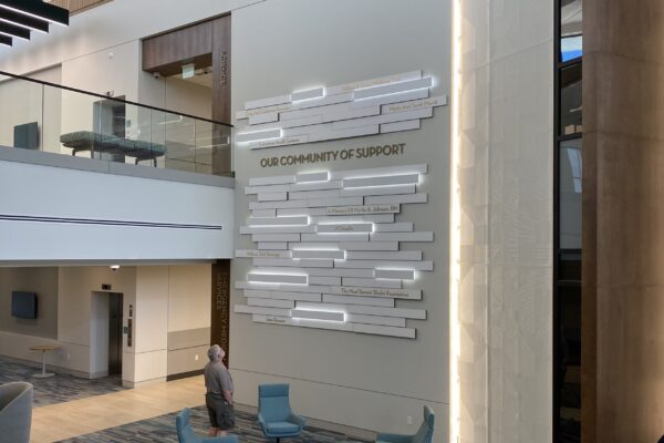 A modern indoor space featuring a high ceiling and contemporary decor. On one wall, there is a large, artistic installation with the text "Our Community of Support." Several chairs and a round table are arranged on the floor.