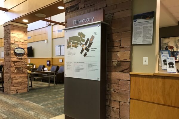 A hospital lobby with stone accents and wooden trim. A large directory sign with a detailed map is prominently displayed. In the background are seating areas with chairs and tables, and a wall-mounted information board is visible on the right.