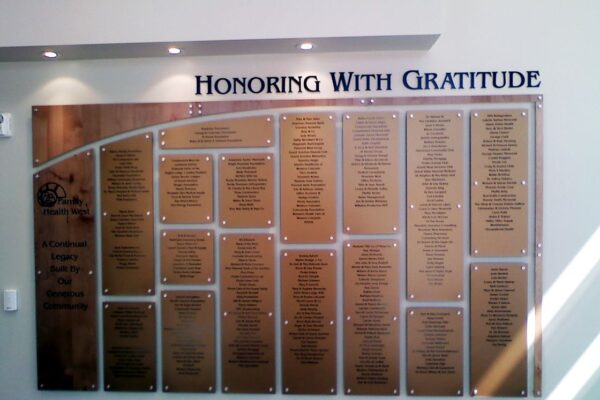 A wall plaque titled "Honoring With Gratitude" displays multiple nameplates with engraved text. The nameplates are arranged in rows and columns on a large board, recognizing individuals for their contributions or achievements.