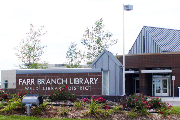 A modern library building with a sign reading "Farr Branch Library - Weld Library District" at the entrance. The building features a mix of brick and metal architecture, surrounded by well-maintained landscaping with flower beds and small trees.