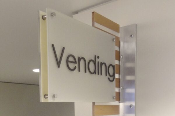 A sign mounted on a wall reads "Vending" in black letters on a frosted glass panel, indicating the location of vending machines. The sign is attached to a metal bracket and is lit from above.