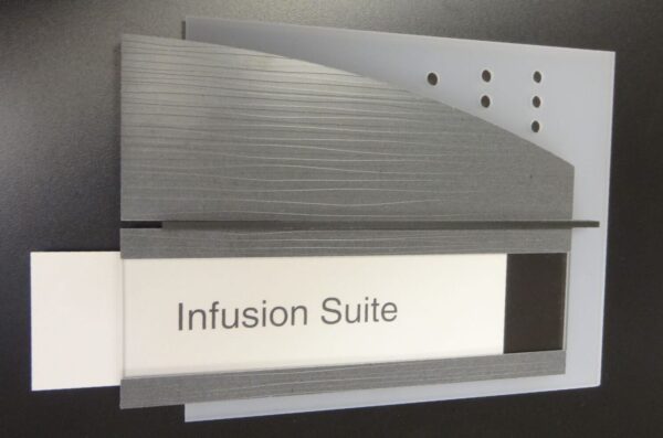 A sign designating the "Infusion Suite" with modern styling. The sign has a grey, textured surface with an angular top, featuring a white panel with the text "Infusion Suite" and several circular cutouts on the grey section. The background is black.