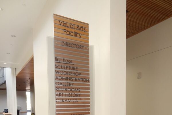 A modern directory sign for the Visual Arts Facility stands in a lobby. It lists various departments including Book Studio, Sculpture, Metalsmithing, Graphic Design, and more. The sign is made of wood with metal accents, next to a hallway with benches.