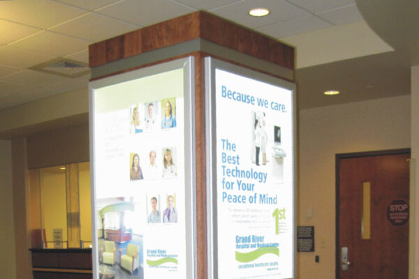 A wooden kiosk stands in a carpeted area with four brightly lit panels displaying various health-related information, images of medical equipment, and texts promoting healthcare services. A reception desk and a door are visible in the background.