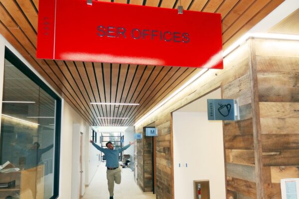 A person mid-jump in a hallway under a red sign that reads "SER Offices." The hallway features wooden paneling on the right side and glass walls on the left. The person is dressed casually and appears to be celebrating or excited.