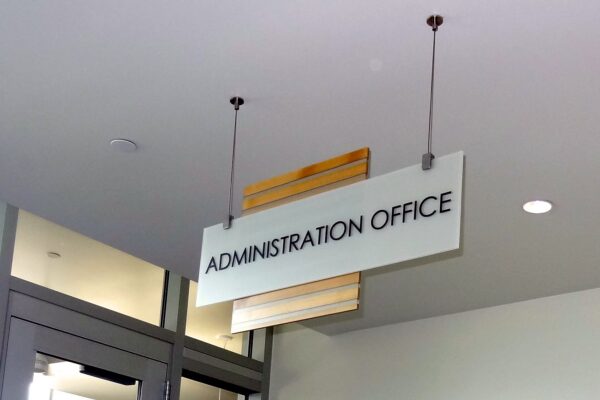 A white rectangular sign with black text reads "ADMINISTRATION OFFICE." The sign is suspended from the ceiling by two wires. The background shows part of a ceiling, walls, and a doorway with glass panels.