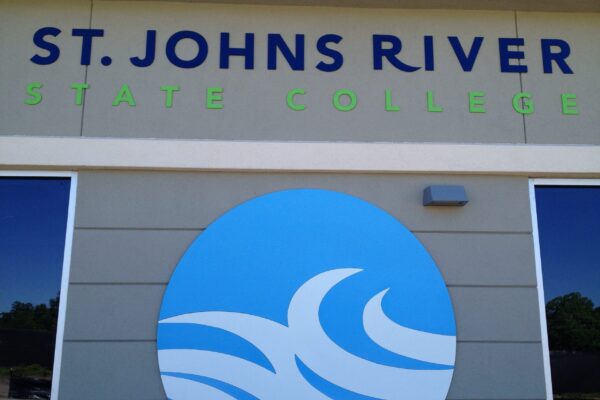 Sign for St. Johns River State College with a blue wave logo against a gray wall. The text "ST. JOHNS RIVER" is in blue, "STATE COLLEGE" is in light green. The background features trees and blue sky partially visible through glass windows on either side.