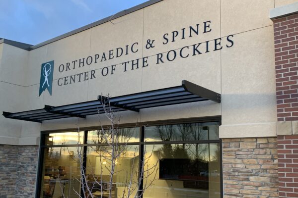 The image shows the exterior of the Orthopaedic & Spine Center of the Rockies building. The signage above the entrance features the center's name in large, dark letters. The building exterior includes stonework, large windows, and a small awning.