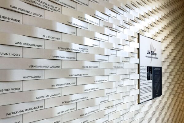 A wall is covered with rows of rectangular white plaques, each bearing different names and messages. To the right, a larger black and white informational plaque is visible. The pattern of the wall creates a textured, wave-like visual effect.