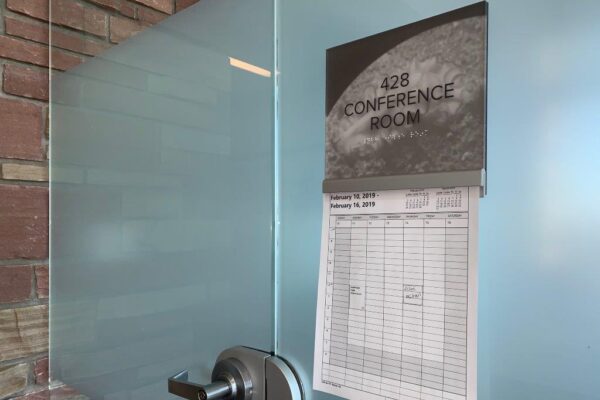 A frosted glass door labeled "Conference Room 428" with a rectangular sign above the handle. A printed schedule with dates and times is attached below the sign. The door handle is silver, and the background wall is made of red brick.