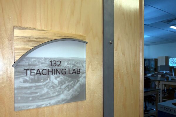 A wooden door with a sign that reads "132 Teaching Lab" in black letters over a black and white photo.