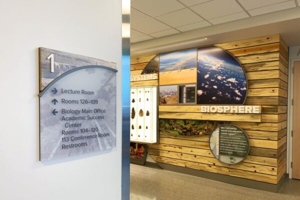 A wall sign on the first floor directs to a lecture room, rooms 126-139, the Biology Main Office, Academic Success Center, rooms 104-120, and a conference room (113). The sign uses a mix of blue and black text with arrows beside each listing.