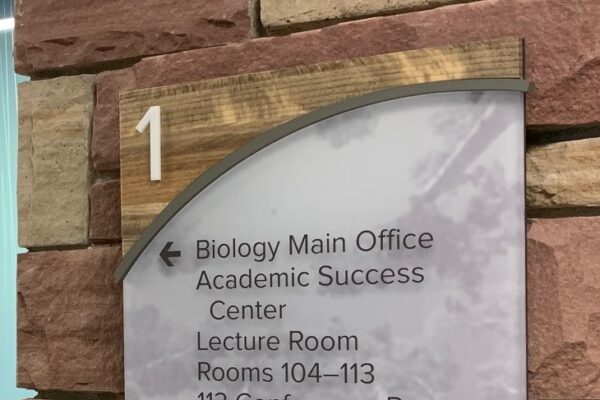 A directional sign mounted on a brick wall with the number 1 at the top. It lists various locations including the Biology Main Office, Academic Success Center, Lecture Room, Rooms 104-113, and the 113 Conference Room, with an arrow pointing left.