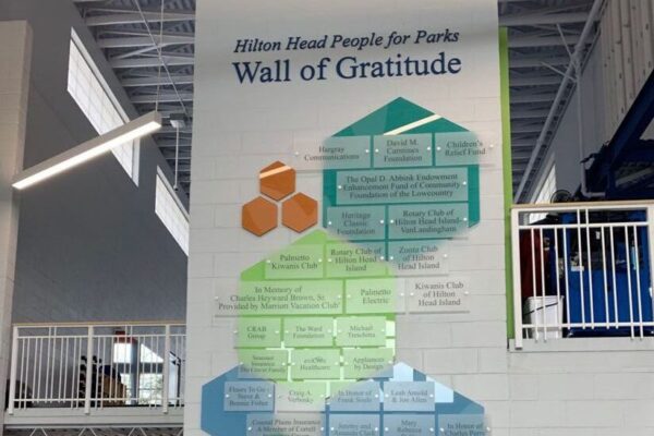 A woman walks up the stairs inside a modern building, passing by a "Wall of Gratitude" display on a large white wall. The display features colorful hexagon shapes containing names and text, signifying recognition and appreciation.