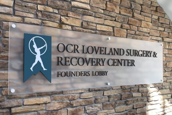 A sign on a brick wall reads "OCR Loveland Surgery & Recovery Center, Founders Lobby." It features a blue emblem with a white silhouette of a person.