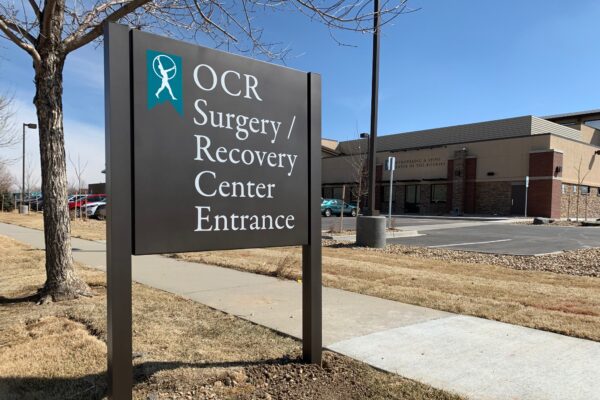 Sign near a sidewalk reads "OCR Surgery / Recovery Center Entrance" with a teal and white logo above the text. A building and parking lot are visible in the background. It is a sunny day with a clear sky.