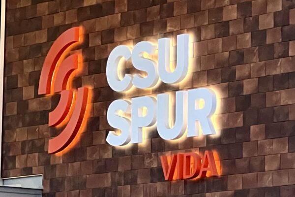 The image shows a wall sign with the text "CSU SPUR VIDA" illuminated. The letters "CSU" and "SPUR" are in white, while the word "VIDA" is in red. The wall is made of brown rectangular tiles, and the letters are outlined with a glowing effect.