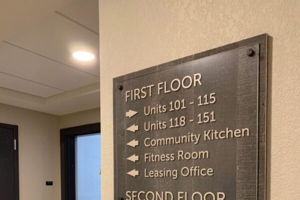 A directional sign mounted on a wall inside a building lists the locations of various rooms and facilities. A hallway and door are visible to the left.
