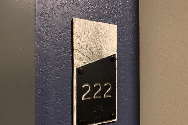 A room number plaque mounted on a textured wall. The plaque has a metallic background with a black section displaying the number "222" in raised text. The wall behind the plaque is painted dark blue.