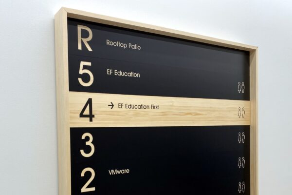 A rectangular directory sign with a wooden frame is mounted on a wall. It lists floors and corresponding occupants: 5 - Rooftop Patio, EF Education; 4 - EF Education First; 3 - VMware; 2 - Fjallraven, Royal Robbins, Wonder Press. The bottom reads "PLATTE FIFTEEN.
