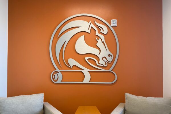 A wall adorned with an orange matte finish displays a stylized horse logo above a scroll. The logo is prominently centered. In front of the wall, two cushioned chairs in a light fabric face each other with a small wooden table placed between them.