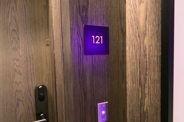 A hotel room door with the number 121 illuminated in purple on a sign above the door handle. The door is made of wood, with a sleek black electronic lock and key card reader below the door number.