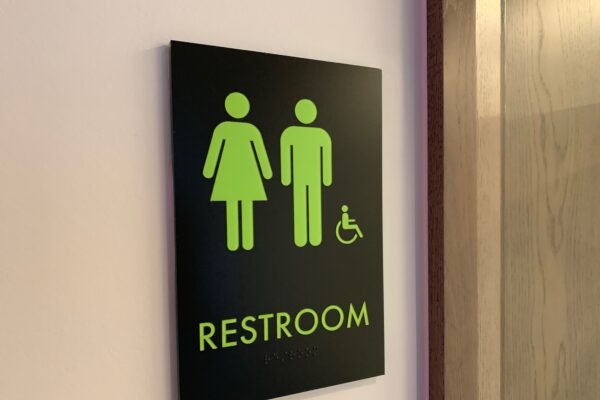 A restroom sign showing green, simplified icons for male, female, and accessible restrooms on a black background. The sign is mounted on a wall next to a wooden door. The word "RESTROOM" is written in green letters beneath the icons.