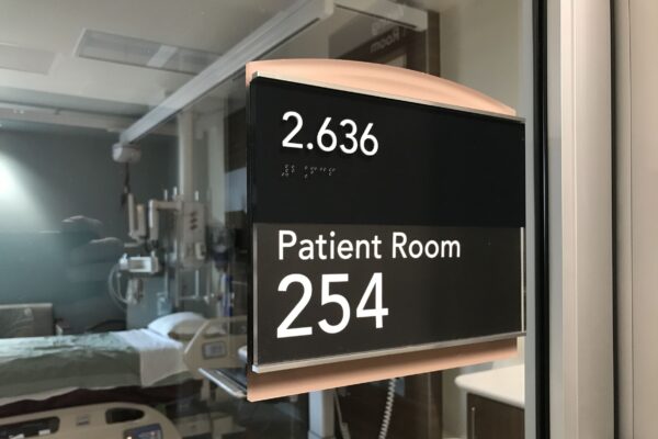 A hospital room sign indicates "2.636" and "Patient Room 254" in white text on a black background. The room, visible through a glass window, contains medical equipment and a bed with white sheets.