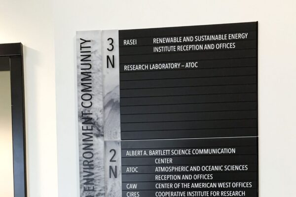 A wall-mounted black and white office directory sign labeled "Sustainability Energy and Environment Community." The sign shows room numbers and corresponding offices or departments. The details of the text are mostly blurry and not fully readable.
