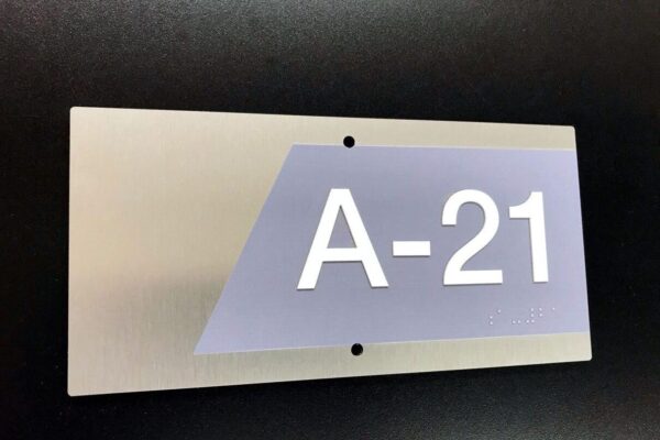 A rectangular metal sign against a dark background displays the characters "A-21" in white. The text is printed on a gray, right-angled trapezoidal section of the sign. Two small holes are visible, one above and one below the text.