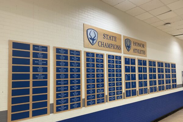 A wall-mounted display board featuring multiple blue plaques with gold trim. The left section is titled "State Champions" and shows numerous names, while the right section is labeled "Honor Athletes" with additional names listed. The wall is white and the floor is wooden.
